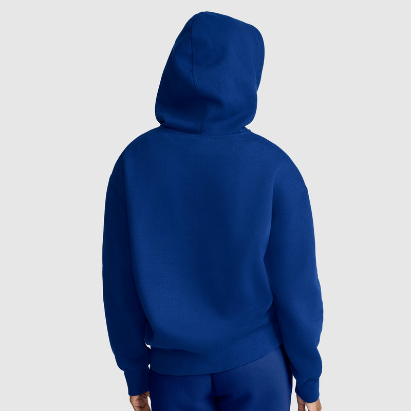 A person wearing a Champion Powerblend Fleece Zip-Up Hoodie, C Logo in blue and matching blue pants is facing away from the camera, standing against a plain light gray background.