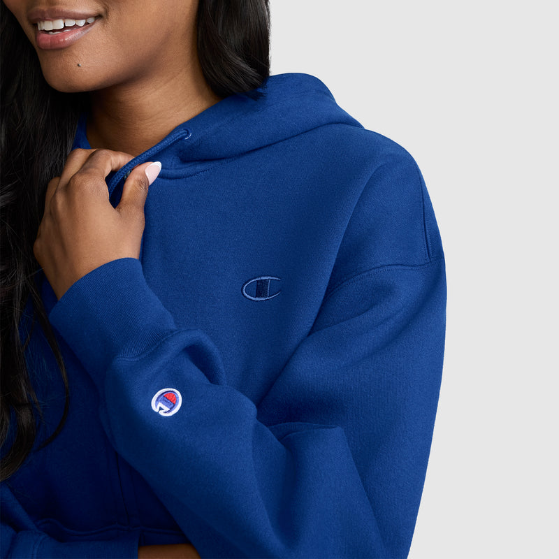 A person wearing a blue Champion Powerblend Fleece Zip-Up Hoodie with a visible C logo on the chest and sleeve, holding the drawstring near their neck.
