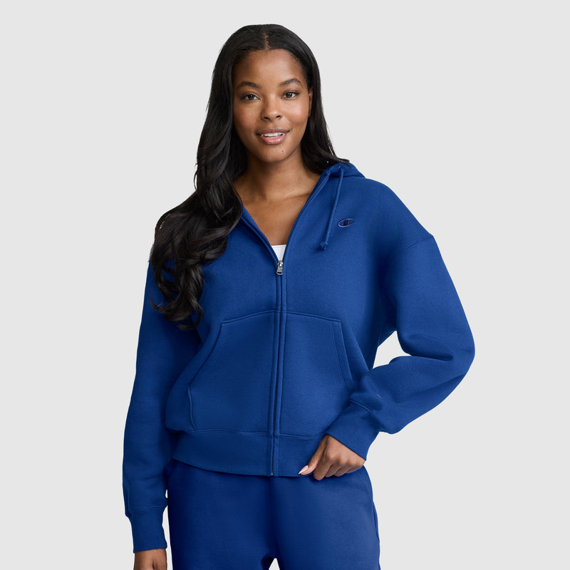 Wearing the Champion Powerblend Fleece Zip-Up Hoodie with C Logo and matching blue pants, a woman stands against a plain light gray background, smiling with one hand in her pocket.