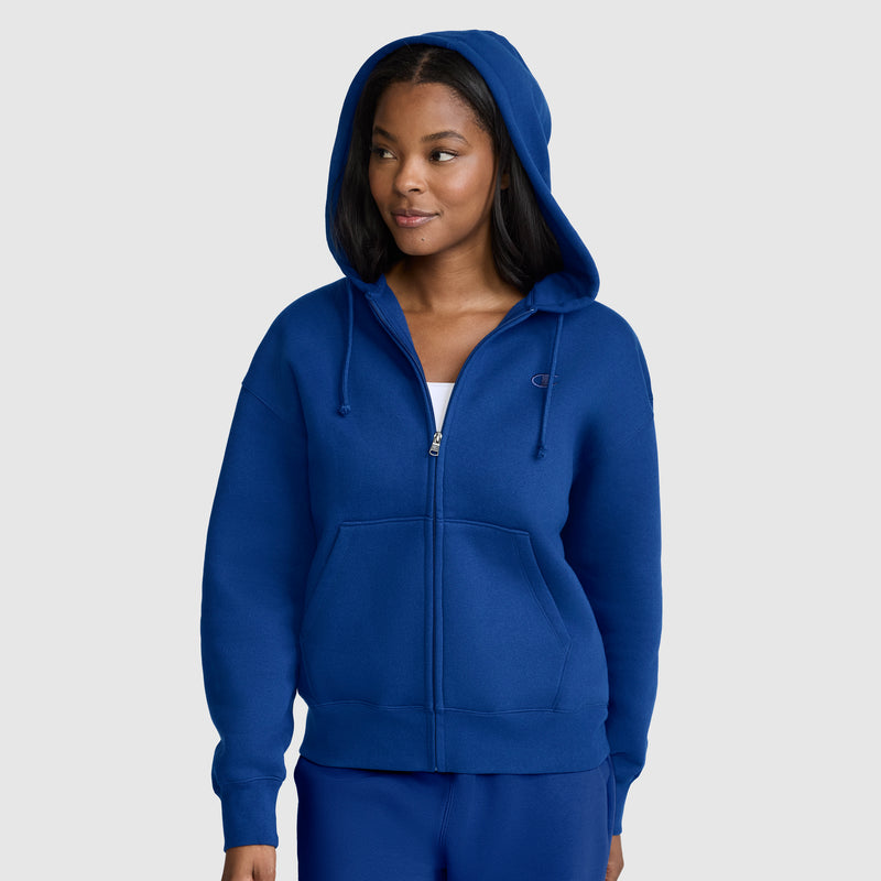 A woman wearing a royal blue Champion Powerblend Fleece Zip-Up Hoodie, C Logo, with the hood up, stands against a plain light gray background.