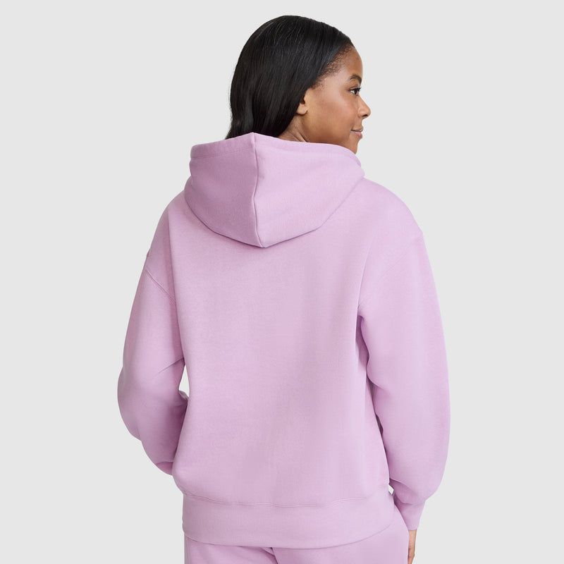 A person with long dark hair wears a light pink Champion Powerblend Fleece Zip-Up Hoodie with the C Logo, along with matching pants, facing away from the camera against a plain light gray background.