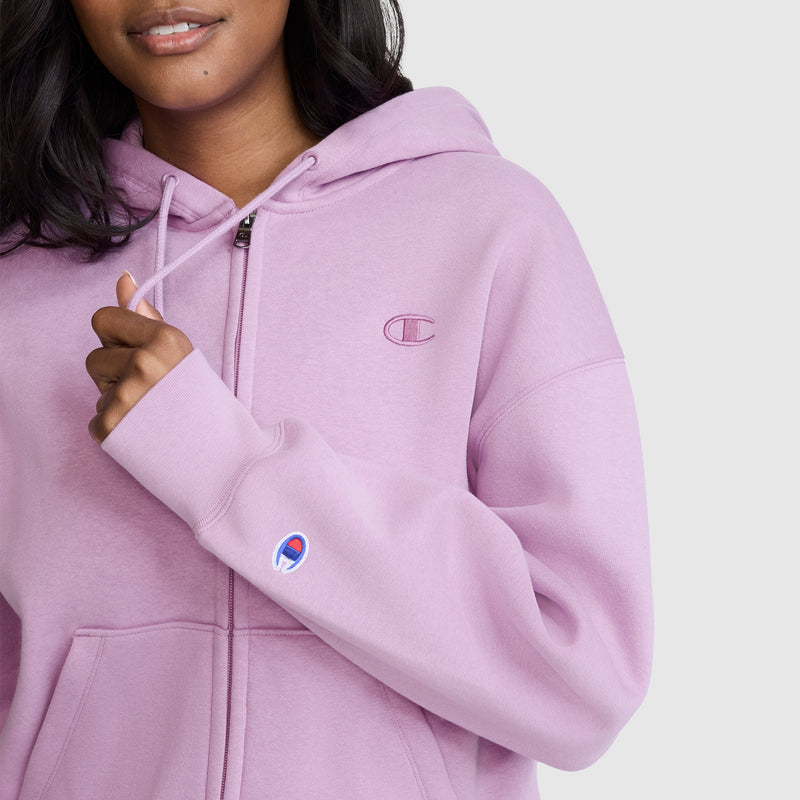 A woman wearing a light purple Champion Powerblend Fleece Zip-Up Hoodie, featuring the C Logo on the sleeve, stands against a plain light gray background.