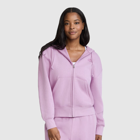 A woman stands against a plain light gray background, smiling softly with her hands by her sides, wearing a Champion Powerblend Fleece Zip-Up Hoodie, C Logo in light purple along with matching sweatpants.