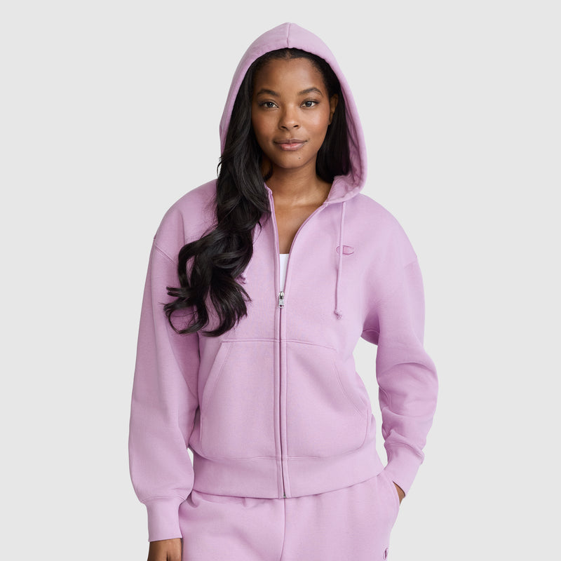 A woman stands facing forward with her hands in the pockets of a light purple Champion Powerblend Fleece Zip-Up Hoodie, featuring a front zipper and a hood pulled over her head. She pairs the hoodie with matching sweatpants.