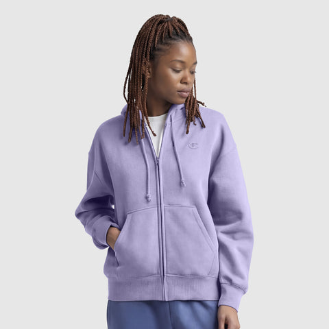 A woman with braided hair wears a light purple Champion Powerblend Fleece Zip-Up Hoodie, C Logo, over a white shirt and blue pants, standing with one hand in her pocket and looking down to the side against a plain background.