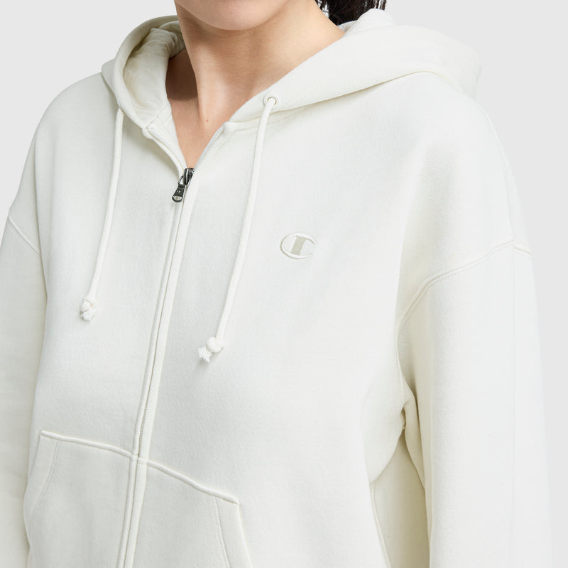 Someone wears a Champion Powerblend Fleece Zip-Up Hoodie, C Logo in off-white with a hood, drawstrings, front pockets, and an embroidered logo on the left chest. The person's face is partially out of frame.