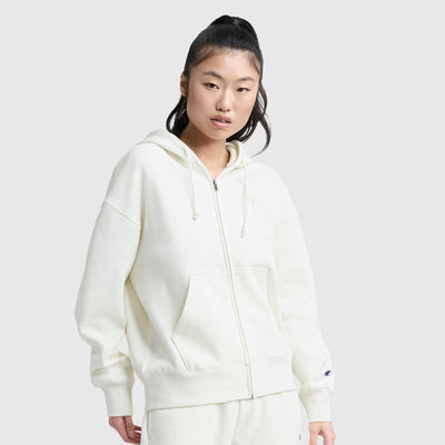 A person with long dark hair in a high ponytail wears a Champion Powerblend Fleece Zip-Up Hoodie, C Logo, paired with matching pants, standing against a plain light gray background.