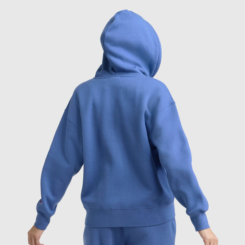 A person is shown from the back, hood up and arms out, wearing a Champion Powerblend Fleece Zip-Up Hoodie, C Logo in blue with matching pants against a plain light gray background.