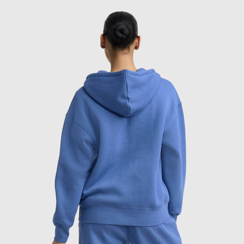 A person with dark hair in a bun is seen from behind, wearing a loose-fitting Champion Powerblend Fleece Zip-Up Hoodie, C Logo in blue with matching blue pants against a plain light gray background.