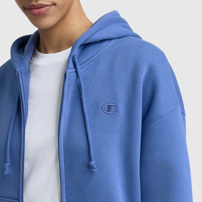 A person wears a blue Champion Powerblend Fleece Zip-Up Hoodie, C Logo, over a white t-shirt. Only the lower half of the face and upper torso are visible, with the embroidered Champion logo on the chest against a plain background.