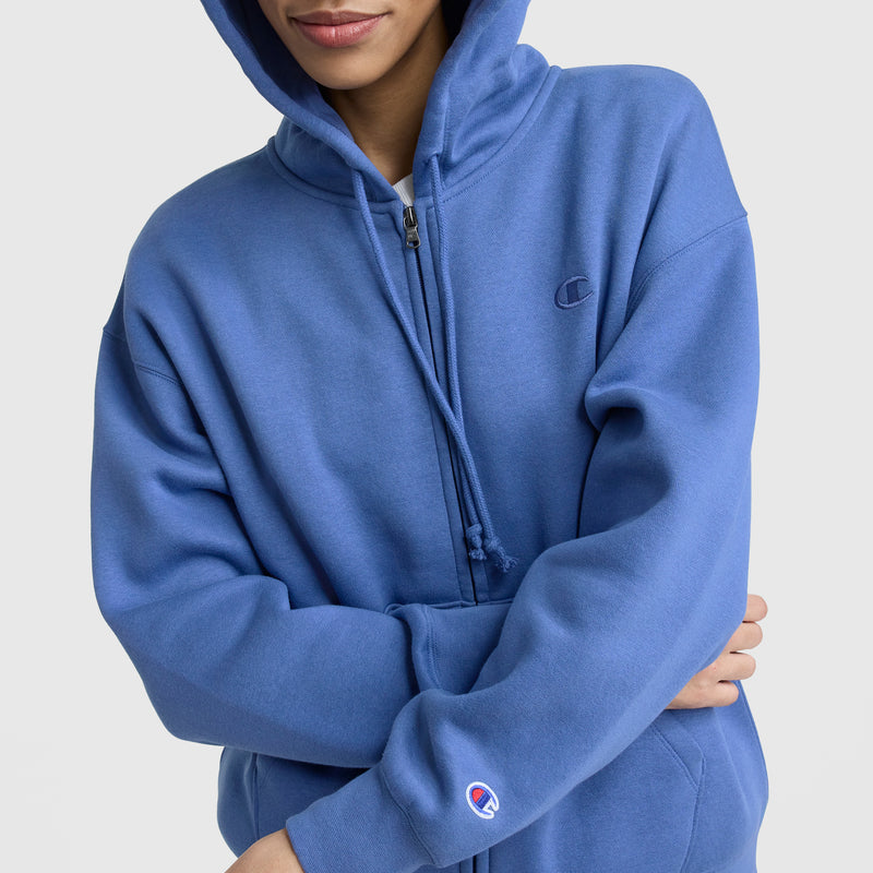 A person wears a blue Champion Powerblend Fleece Zip-Up Hoodie, C Logo, with the hood up and arms crossed. The Champion logo is visible on the chest and sleeve; part of their face is out of frame.