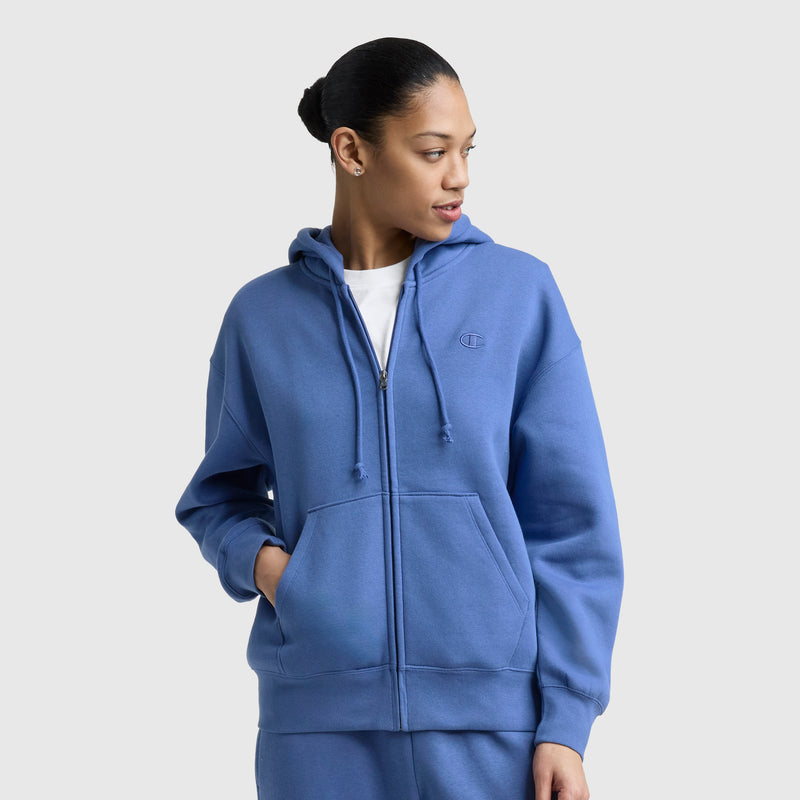 A woman with dark hair pulled back, hands in pockets, models the Champion Powerblend Fleece Zip-Up Hoodie, C Logo in blue against a plain light gray background, looking to her left.
