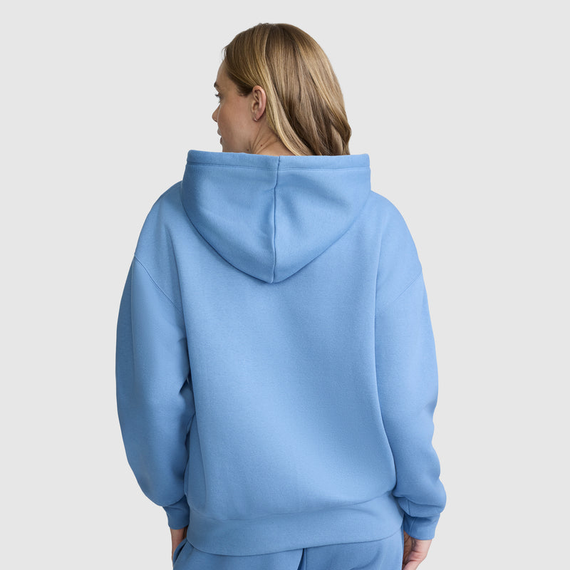A person with shoulder-length light brown hair stands with their back to the camera against a plain light gray background, wearing the Champion Powerblend Fleece Zip-Up Hoodie, C Logo in light blue along with matching pants.