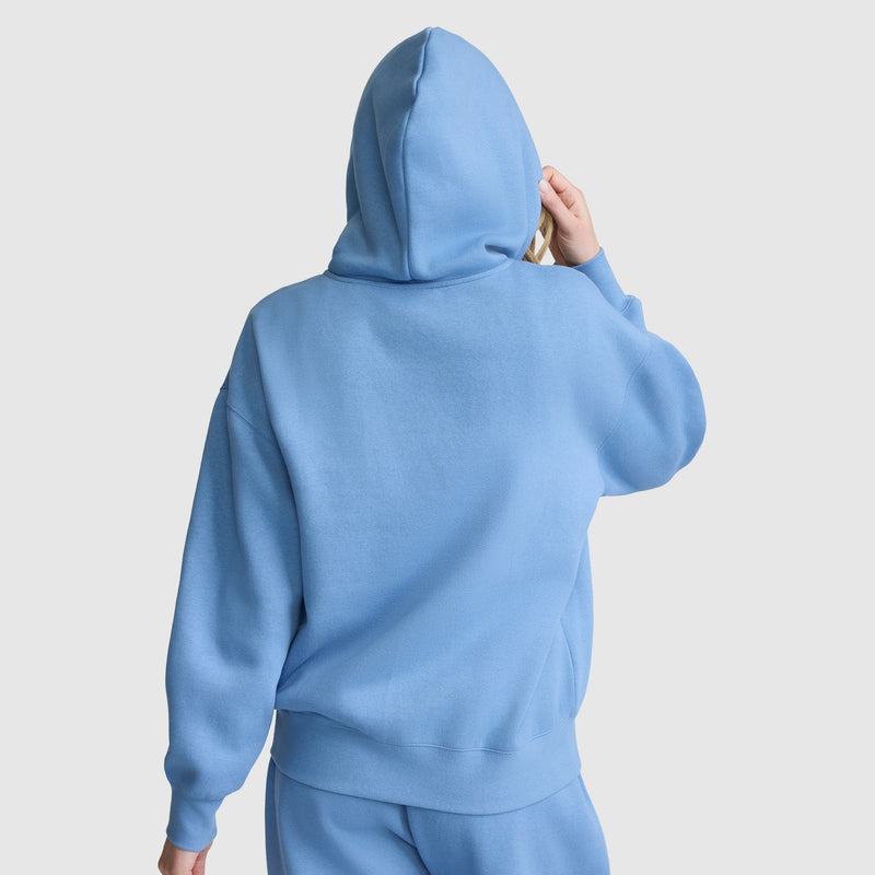 A person wearing a loose-fitting, light blue Champion Powerblend Fleece Zip-Up Hoodie, C Logo, along with matching pants is facing away from the camera with the hood up and one hand touching the side of the hood. The background is plain white.