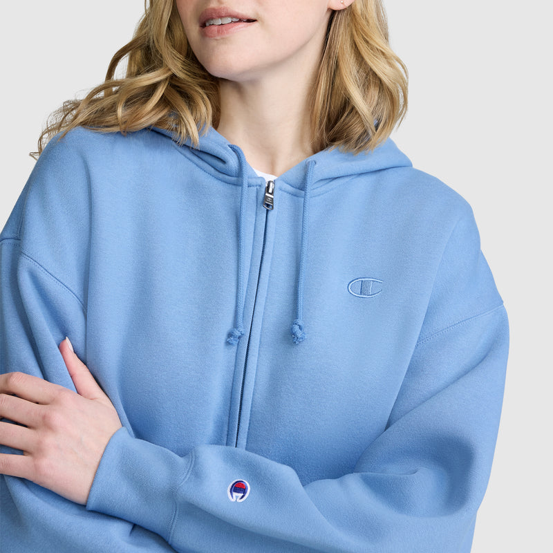 A woman in a light blue Champion Powerblend Fleece Zip-Up Hoodie, C Logo stands with her arms crossed, displaying the embroidered chest and sleeve logos against a plain gray background. Made from Champion’s signature Powerblend fleece, this women’s athletic hoodie delivers both comfort and sporty style.