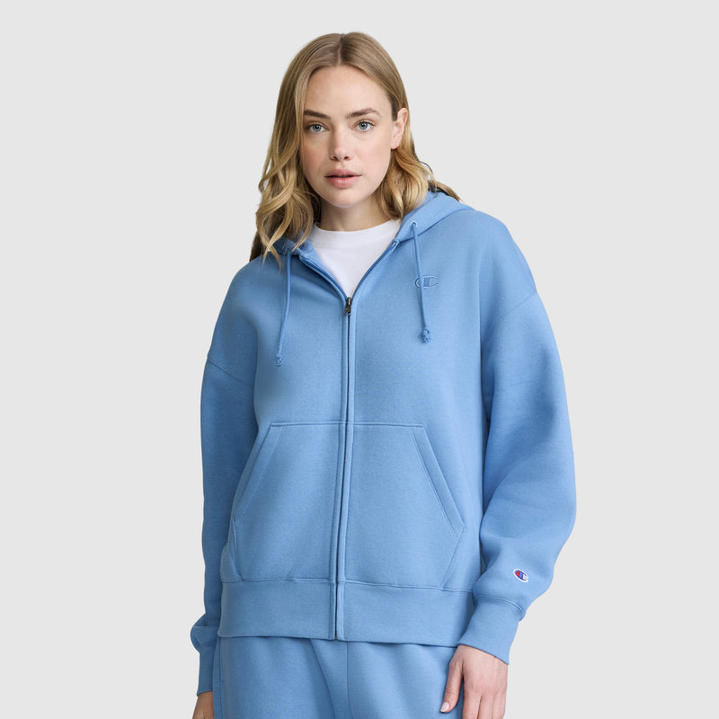 A woman with blonde hair wears a light blue Champion Powerblend Fleece Zip-Up Hoodie, C Logo, along with matching sweatpants, standing against a plain white background.