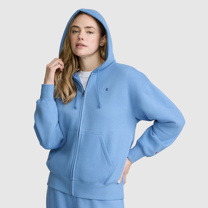 A woman wearing the Champion Powerblend Fleece Zip-Up Hoodie, C Logo, with the hood up and paired with matching sweatpants, stands against a plain light gray background.