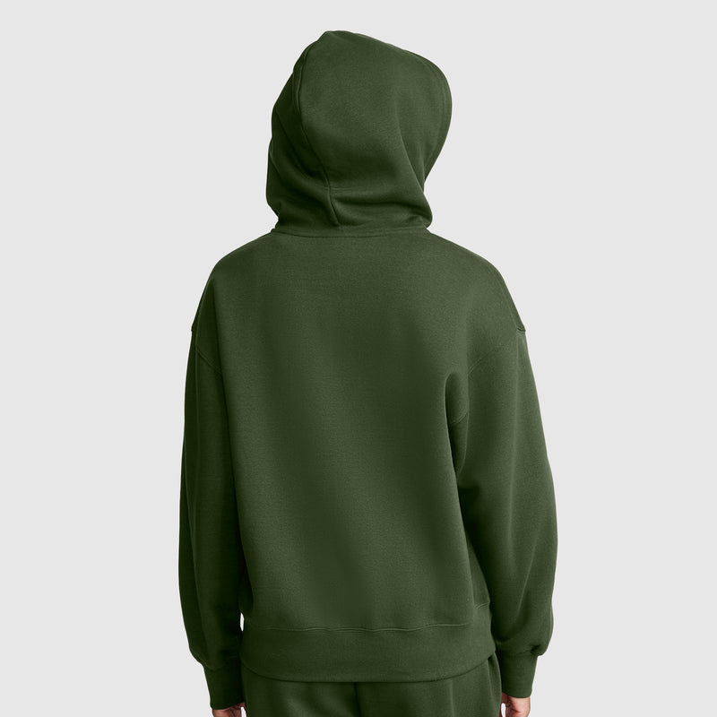 A person wearing a dark green Champion Powerblend Fleece Zip-Up Hoodie, C Logo, with the hood up faces away from the camera, set against a plain light gray background.