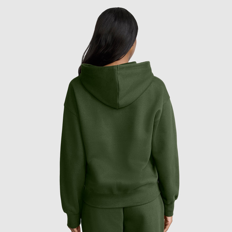 A person with long dark hair is wearing a Champion Powerblend Fleece Zip-Up Hoodie, C Logo, in dark green along with matching pants, facing away from the camera against a plain light background.