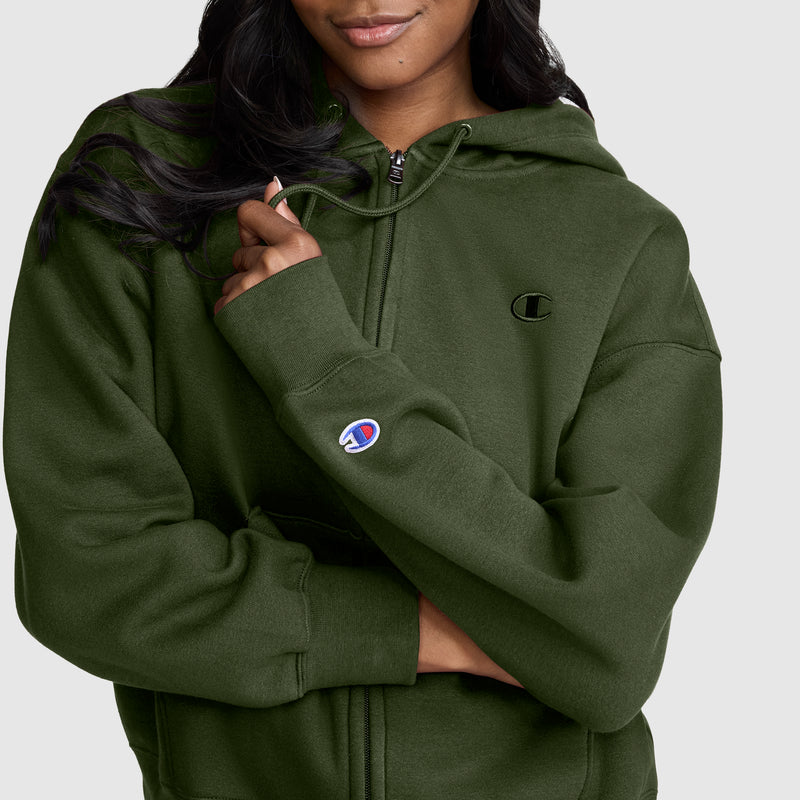 A woman wearing the Champion Powerblend Fleece Zip-Up Hoodie, featuring the signature C logo on the chest and sleeve, crosses her arms and smiles slightly against a plain background. This athletic zip-up hoodie combines comfort and style for everyday wear.