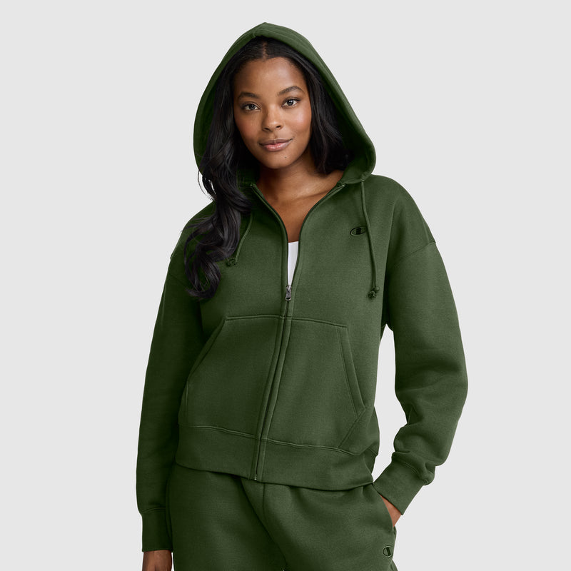 Wearing a dark green Champion Powerblend Fleece Zip-Up Hoodie with the C Logo, a woman stands with one hand in her pocket and smiles slightly against a plain light gray background.