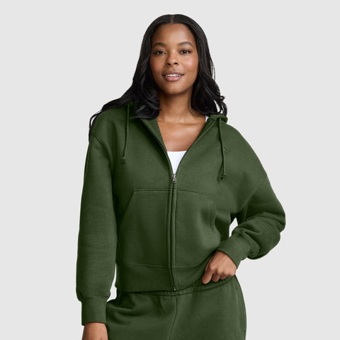 A woman wearing a dark green Champion Powerblend Fleece Zip-Up Hoodie with a C logo and matching sweatpants stands against a plain light gray background, smiling slightly with one hand on her hip.