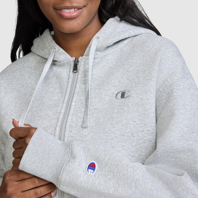 A person wears a light gray Champion Cropped Zip-Up Hoodie, featuring a small C logo on the chest and a blue-and-red embroidered logo on the sleeve. Only the lower face and upper body are visible.