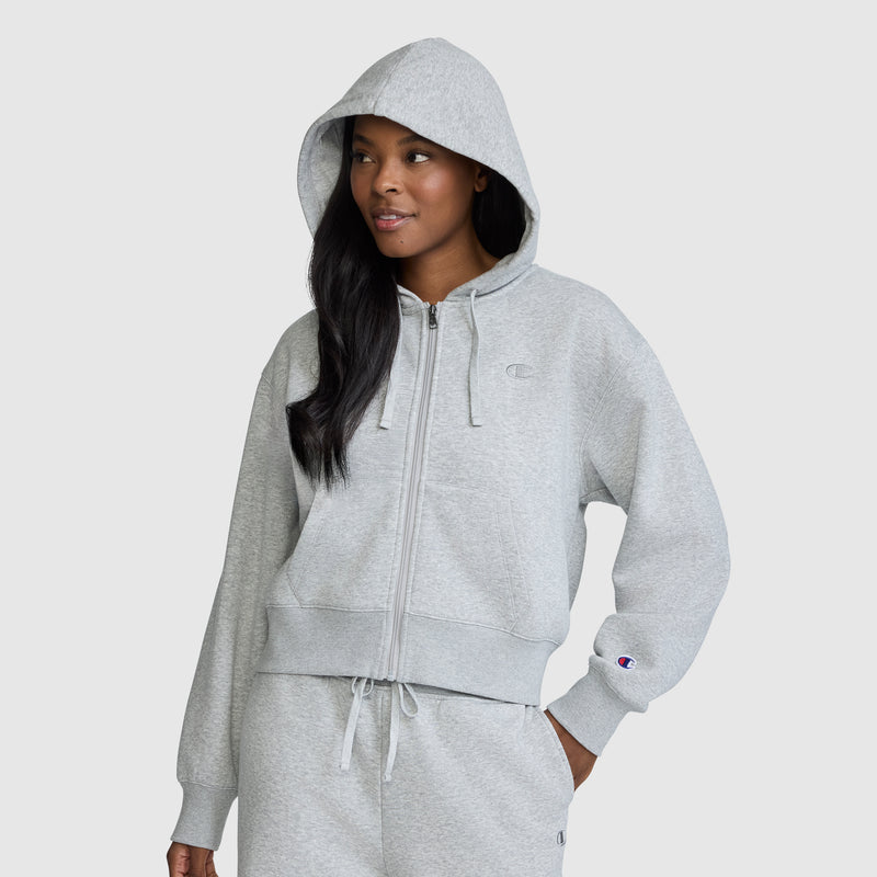 A woman models the Champion Cropped Zip-Up Hoodie in light gray with the hood up, paired with matching sweatpants, standing against a plain light background and looking to the side with a slight smile.