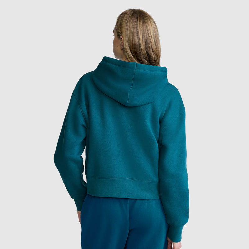 A person with light brown hair stands with their back to the camera against a plain light gray background, wearing a teal Champion Cropped Zip-Up Hoodie and matching pants.
