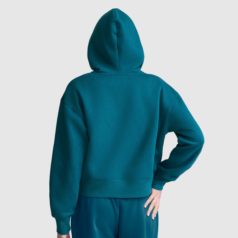 A person stands facing away, wearing a teal Champion Cropped Zip-Up Hoodie with matching pants. One hand rests on their hip, and the background is plain and light-colored.