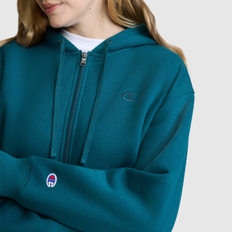 A woman wearing a teal Champion Cropped Zip-Up Hoodie, featuring the iconic Champion logo on the chest and sleeve, stands with arms crossed against a plain light gray background. This cozy fleece hoodie combines style and comfort, making it perfect for any casual outfit.