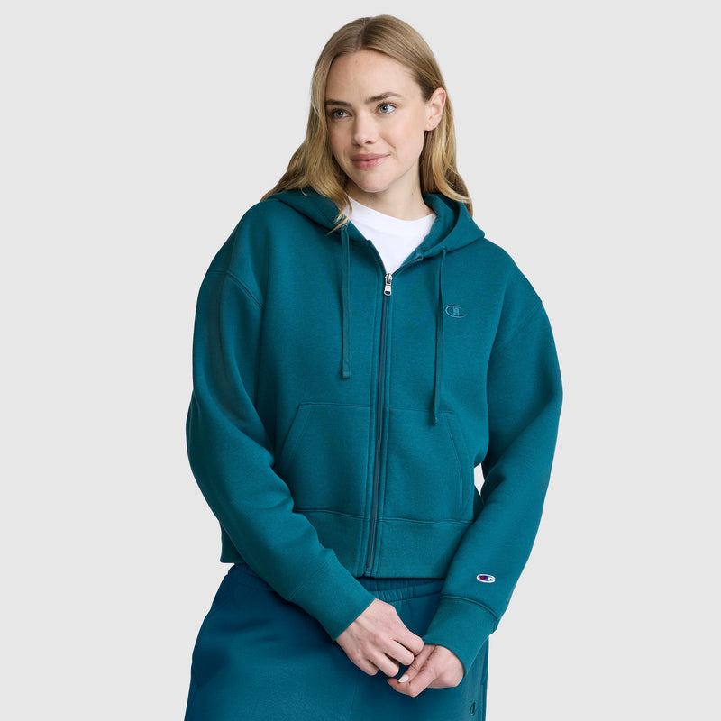 A woman with blonde hair, wearing a teal Champion Cropped Zip-Up Hoodie and matching pants, stands against a plain light gray background, looking slightly to the side with a soft smile.