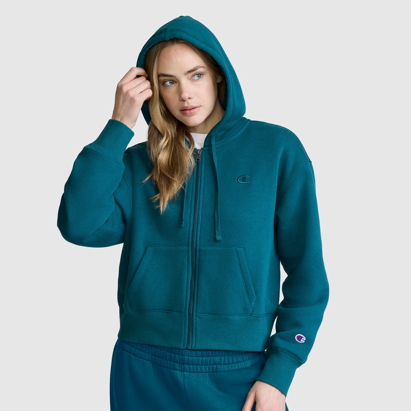 A woman wearing the Champion Cropped Zip-Up Hoodie in teal, paired with matching pants, stands against a plain background with her right hand touching her hair and the hood pulled up over her head.