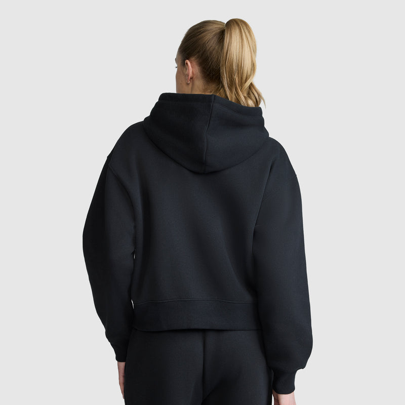 A person with light brown hair in a ponytail is seen from behind, standing against a plain light gray background and wearing the Champion Cropped Zip-Up Hoodie in black along with black pants.