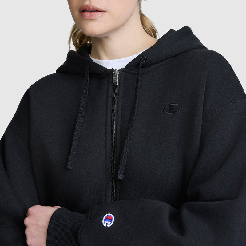 A person wearing a black Champion Cropped Zip-Up Hoodie with a small embroidered logo on the chest and sleeve stands against a plain light background. This cozy fleece hoodie combines comfort and style with its zip-up design.