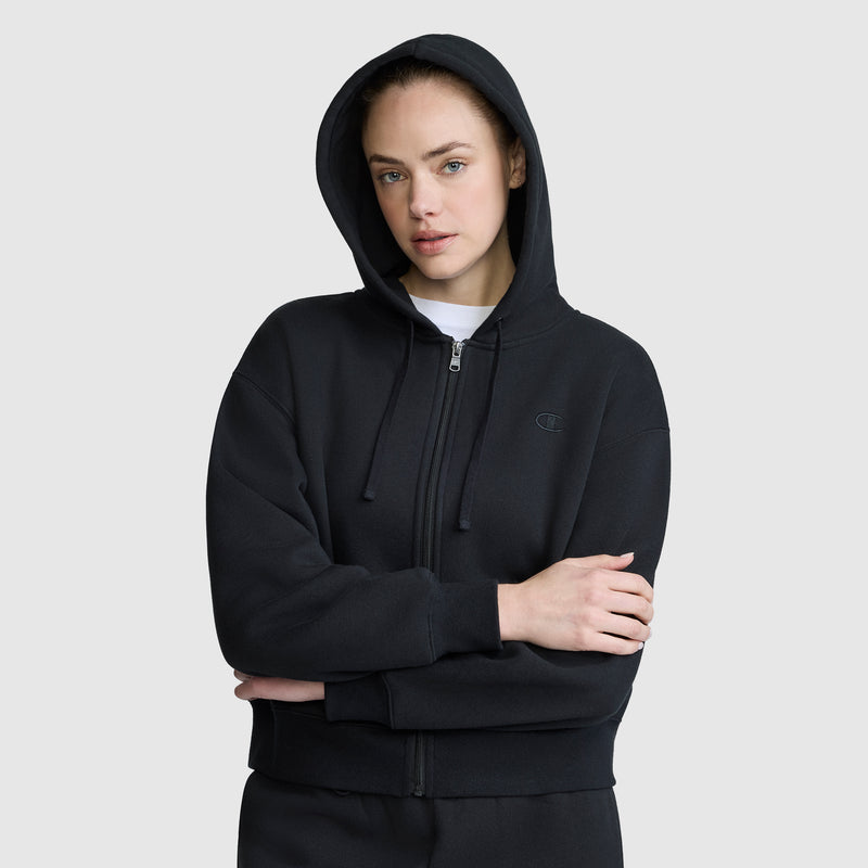 A person wearing a black Champion Cropped Zip-Up Hoodie with the hood up, standing against a plain light gray background, arms crossed and looking at the camera with a neutral expression.