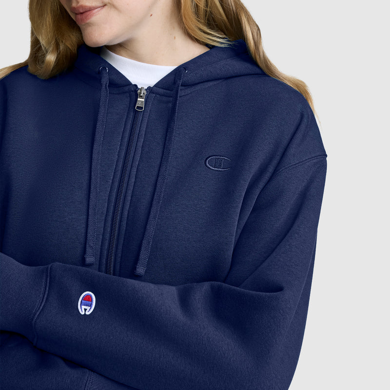 A person wearing a navy blue Champion Cropped Zip-Up Hoodie, featuring the iconic logo on the sleeve and chest, stands against a plain light background.