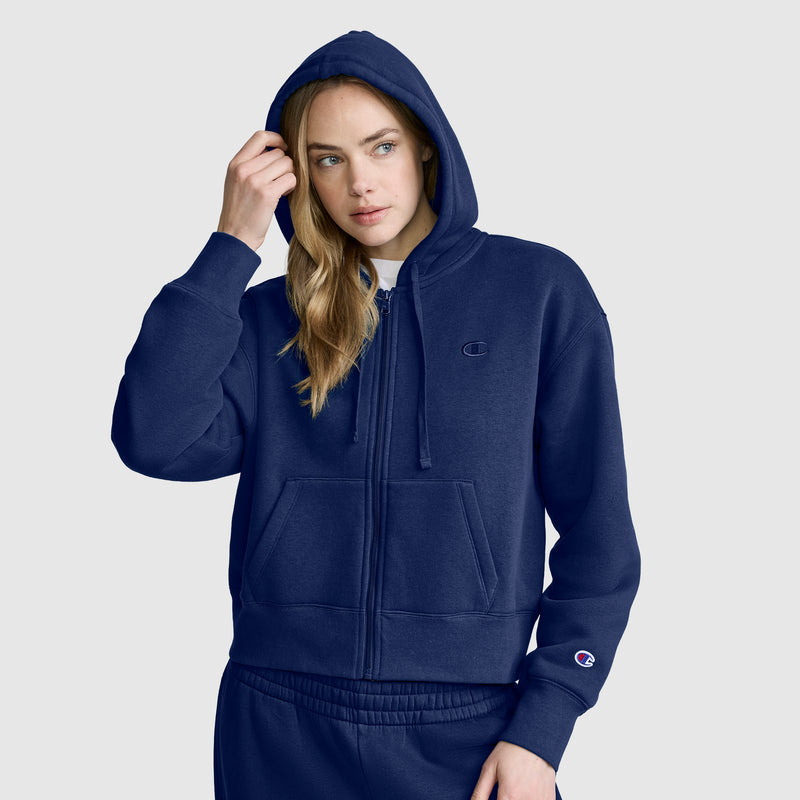 A person wearing a Champion Cropped Zip-Up Hoodie in navy blue with the hood up and matching sweatpants stands against a plain light gray background.
