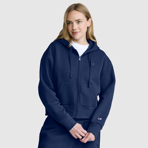A woman wearing a navy blue Champion Cropped Zip-Up Hoodie and matching sweatpants stands against a light gray background, looking slightly to the side and smiling softly.