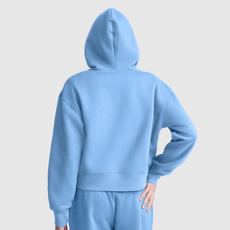 A person is seen from behind, wearing a plain, light blue Champion Cropped Zip-Up Hoodie and matching sweatpants against a neutral background. Their right hand rests on their hip.