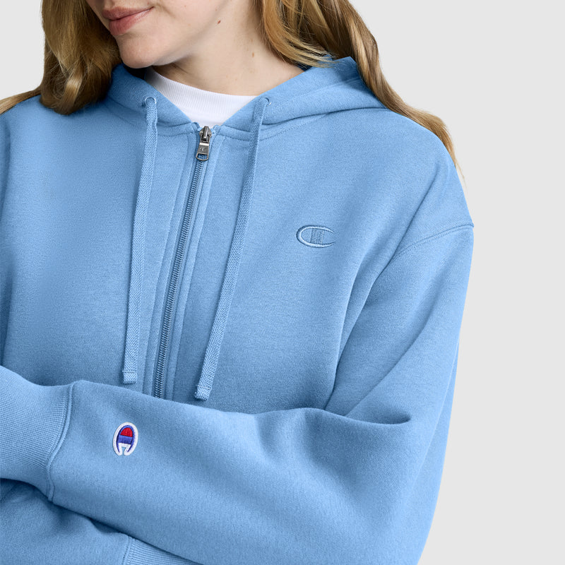 A woman wearing a light blue Champion Cropped Zip-Up Hoodie with drawstrings stands against a plain light background, her arms crossed and face partially visible. This hoodie offers cozy comfort and casual style with its classic zip-up design.