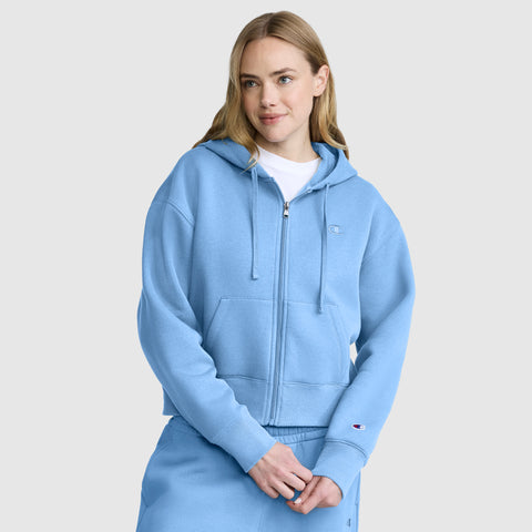 A woman wearing a light blue Champion Cropped Zip-Up Hoodie and matching sweatpants stands against a plain light gray background, smiling slightly with her hands clasped in front of her.