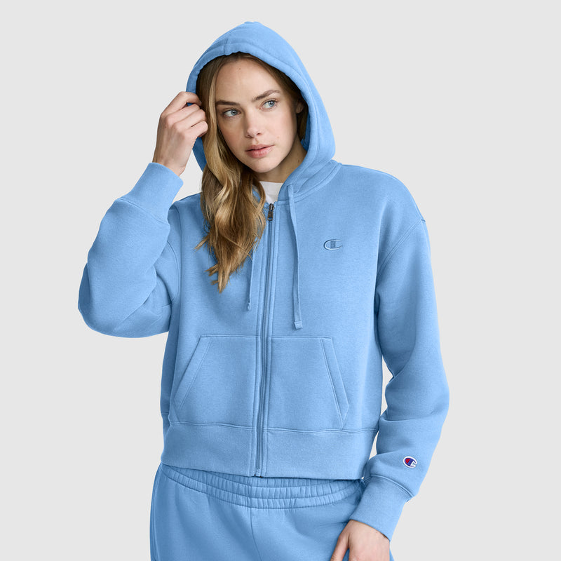 A woman wearing a light blue Champion Cropped Zip-Up Hoodie and matching sweatpants stands against a plain background, her left hand touching her hood and her right hand in her pocket.