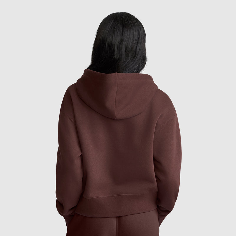 A person with long dark hair is wearing a brown Champion Cropped Zip-Up Hoodie and matching pants, standing with their back facing the camera against a plain light gray background.