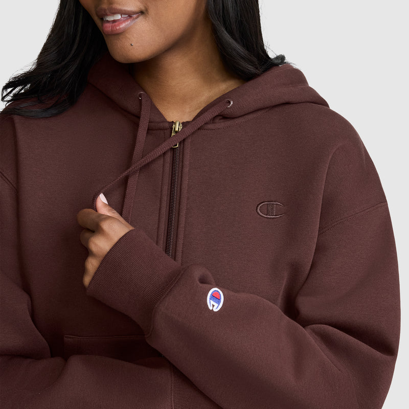 A woman wears the Champion Cropped Zip-Up Hoodie in brown, holding one drawstring. The zip-up hoodie displays the Champion logo on both her sleeve and chest. She smiles softly, shown from the shoulders up against a plain background.