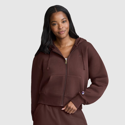 A woman wearing an olive green Champion zip-up hoodie and matching sweatpants against a light gray studio background.