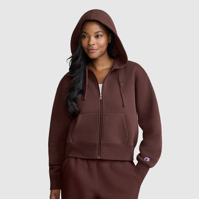 A woman wearing the Champion Cropped Zip-Up Hoodie in brown with the hood up, paired with matching sweatpants, stands against a plain light gray background.