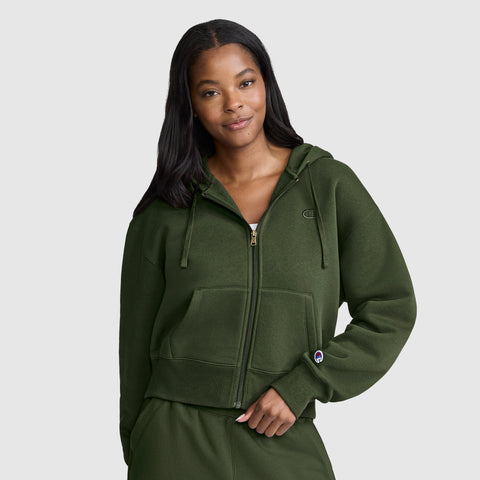 A woman with long dark hair wears a green Champion Cropped Zip-Up Hoodie and matching pants. She stands against a plain gray background, smiling softly with one hand in her pocket.