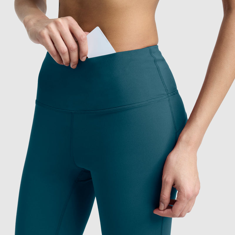 Close-up of a person wearing Champion Soft Touch Flare Leggings, Anti Odor, featuring the C Logo for women, placing a white card into a hidden waistband pocket. The background is plain and light gray.