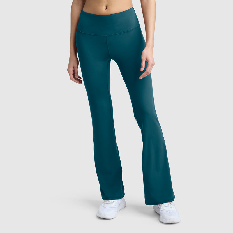 A person models the Champion Soft Touch Flare Leggings featuring anti-odor technology and a C logo in teal, paired with white sneakers against a plain light gray background. Only the lower torso and legs are shown.